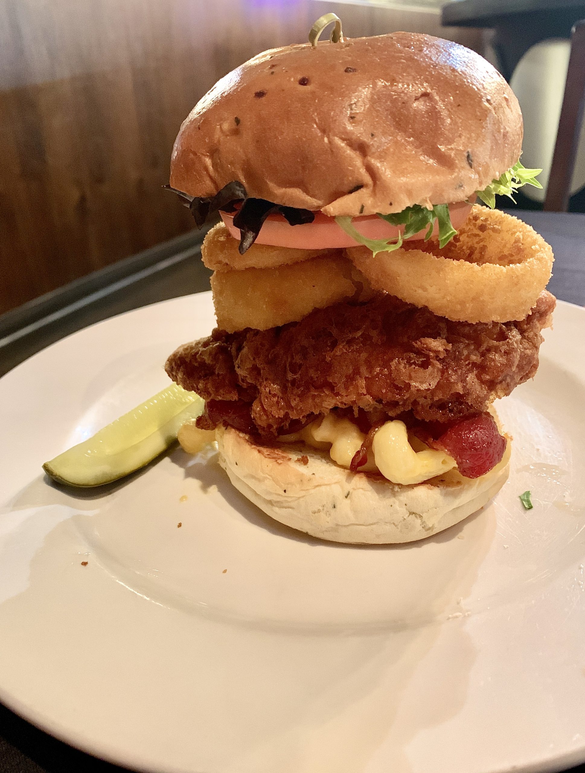 a very tall stacked burger loaded with mac and cheese, louisiana breaded chicken, onion rings, lettuce and tomato on a white plate.