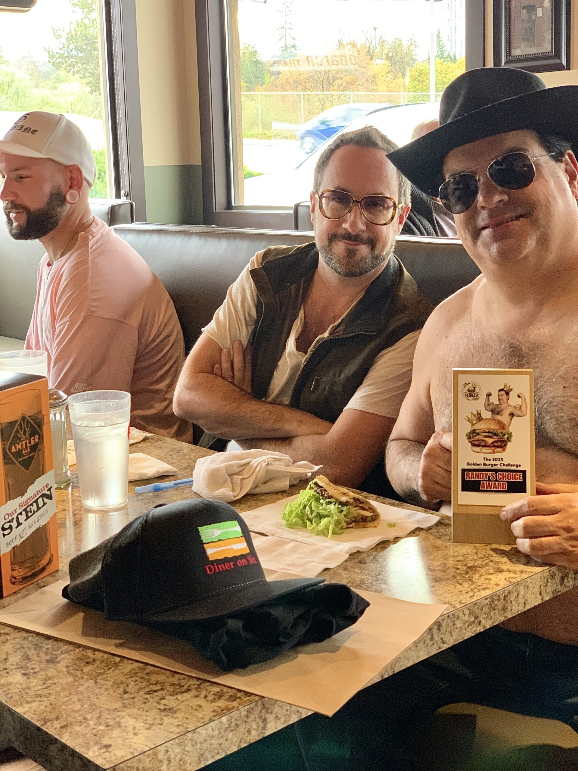 a shirtless Randy from Trailer Park boys and his media team sit down at a restaurant booth to enjoy a taco burger. Randy has a cowboy hat and sunglasses on