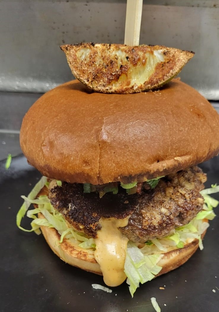 a burger on a black plate with a beef patty, sauce pouring out, lettuce and a bun topped with a lime wedge.