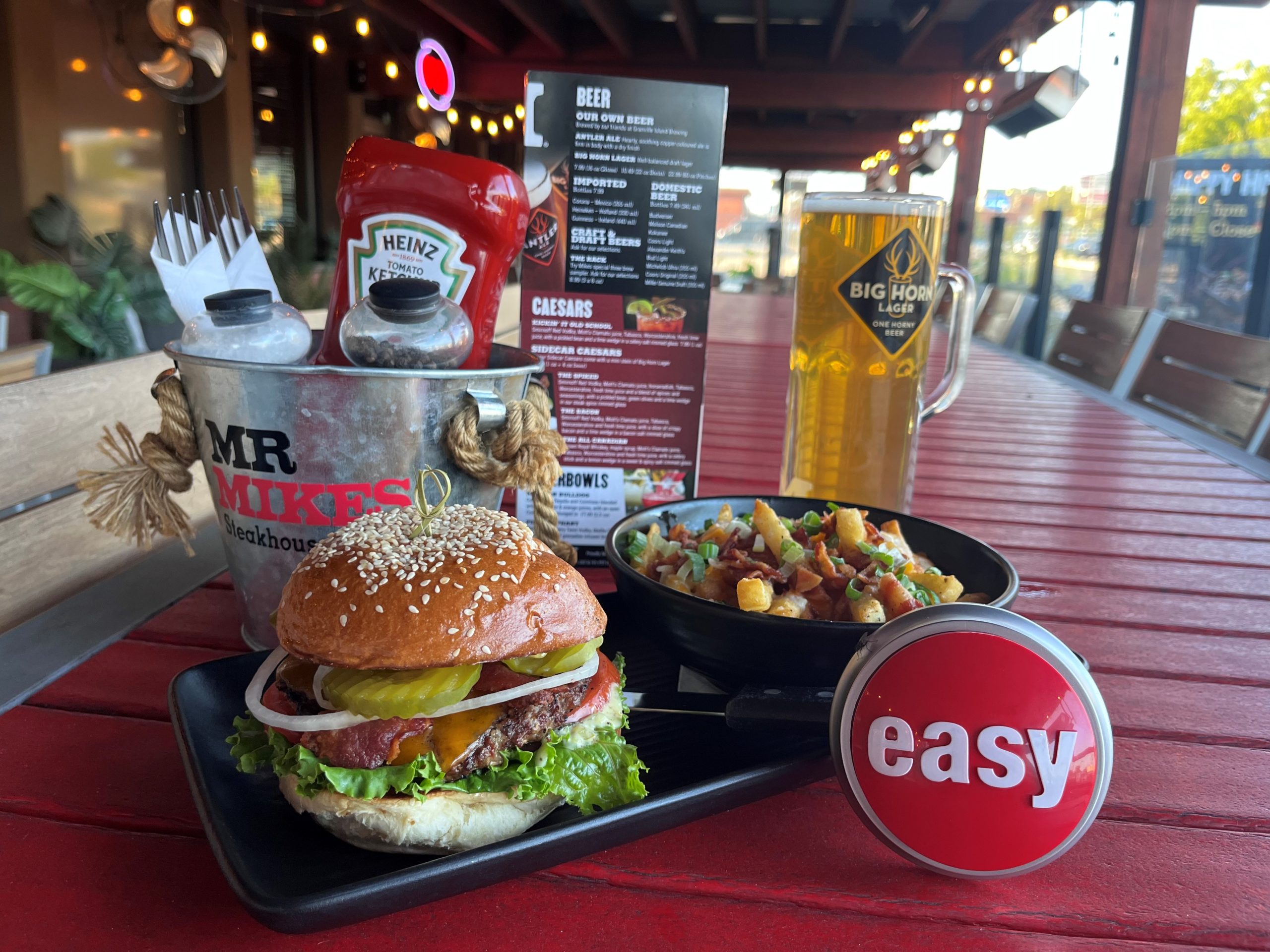 A burger on a black plate with fries on a red patio table. Next to the plate is a Staples "easy" button, some condiments, and a beer in a tall glass