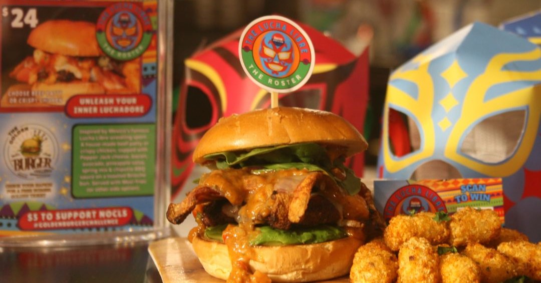 A burger with sauce dripping down on a plate with tater tots beside it. jMexican wrestling masks and items in background