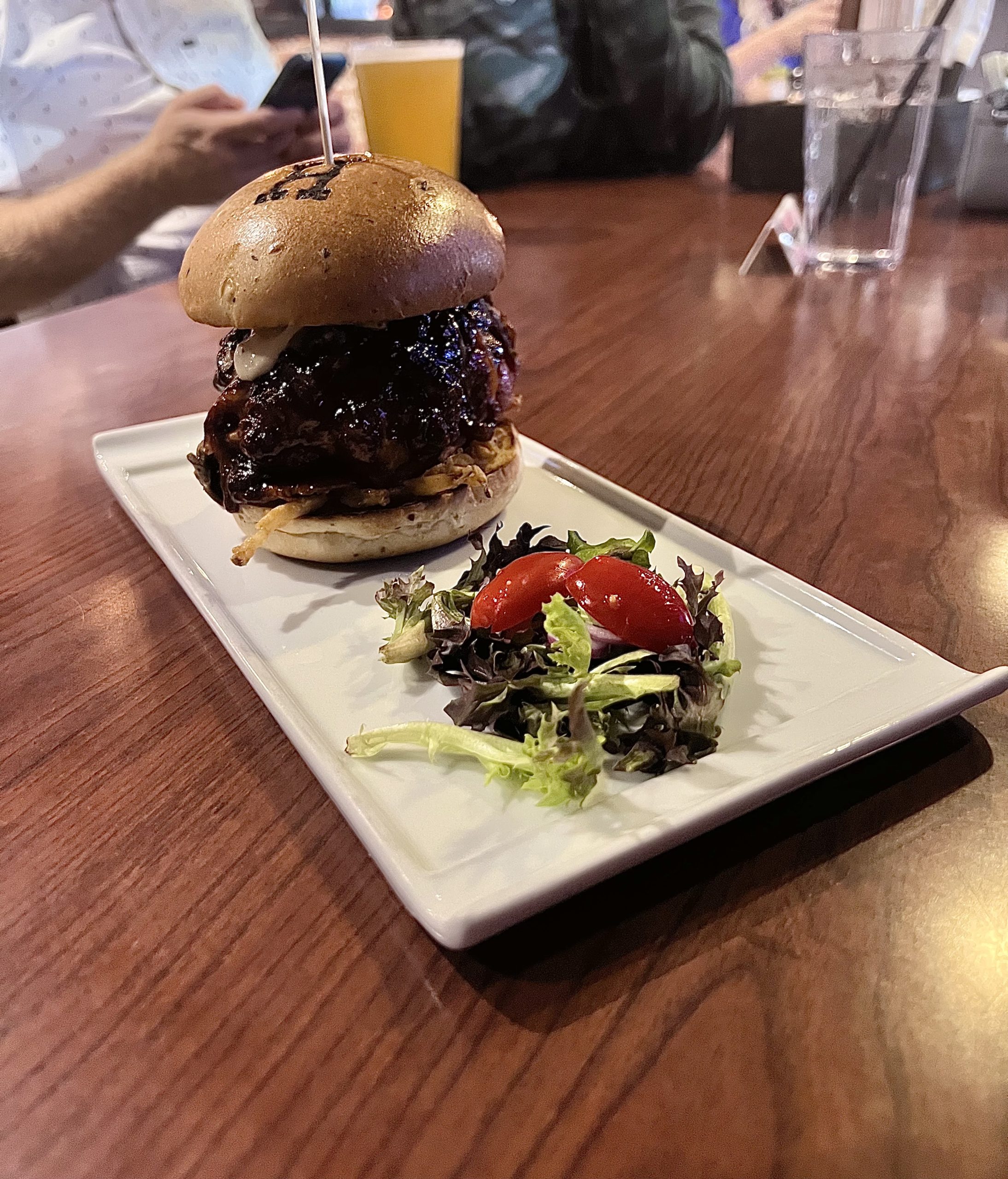 A very tall burger on a long white plate on a table with a small salad on the side