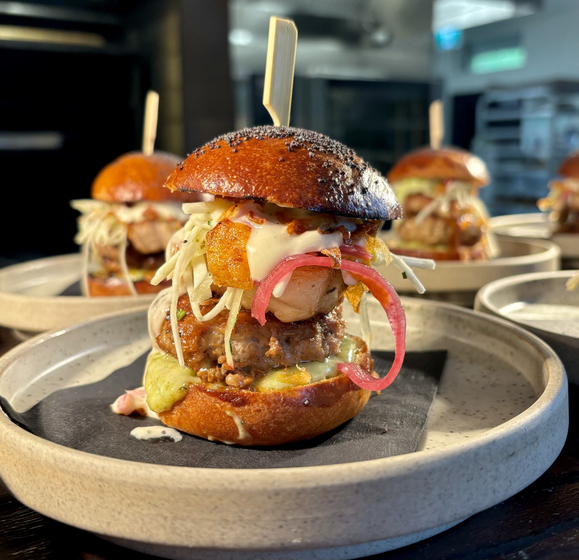 A burger featuring a juicy pork patty topped with succulent, apple cider BBQ-glazed pork belly. Finished with a drizzle of creamy cheese sauce and tangy pickled onions. All served on a soft, buttery brioche bun.