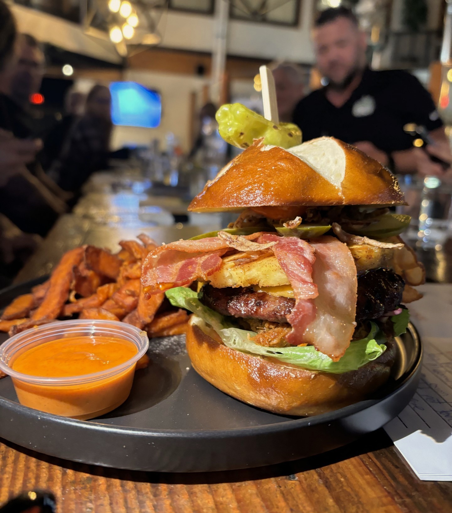 A buger that is stacked very tall with a beef patty, bacon, pineapple, and lettuce. A side of yam fries and dipping sauce on the plate.