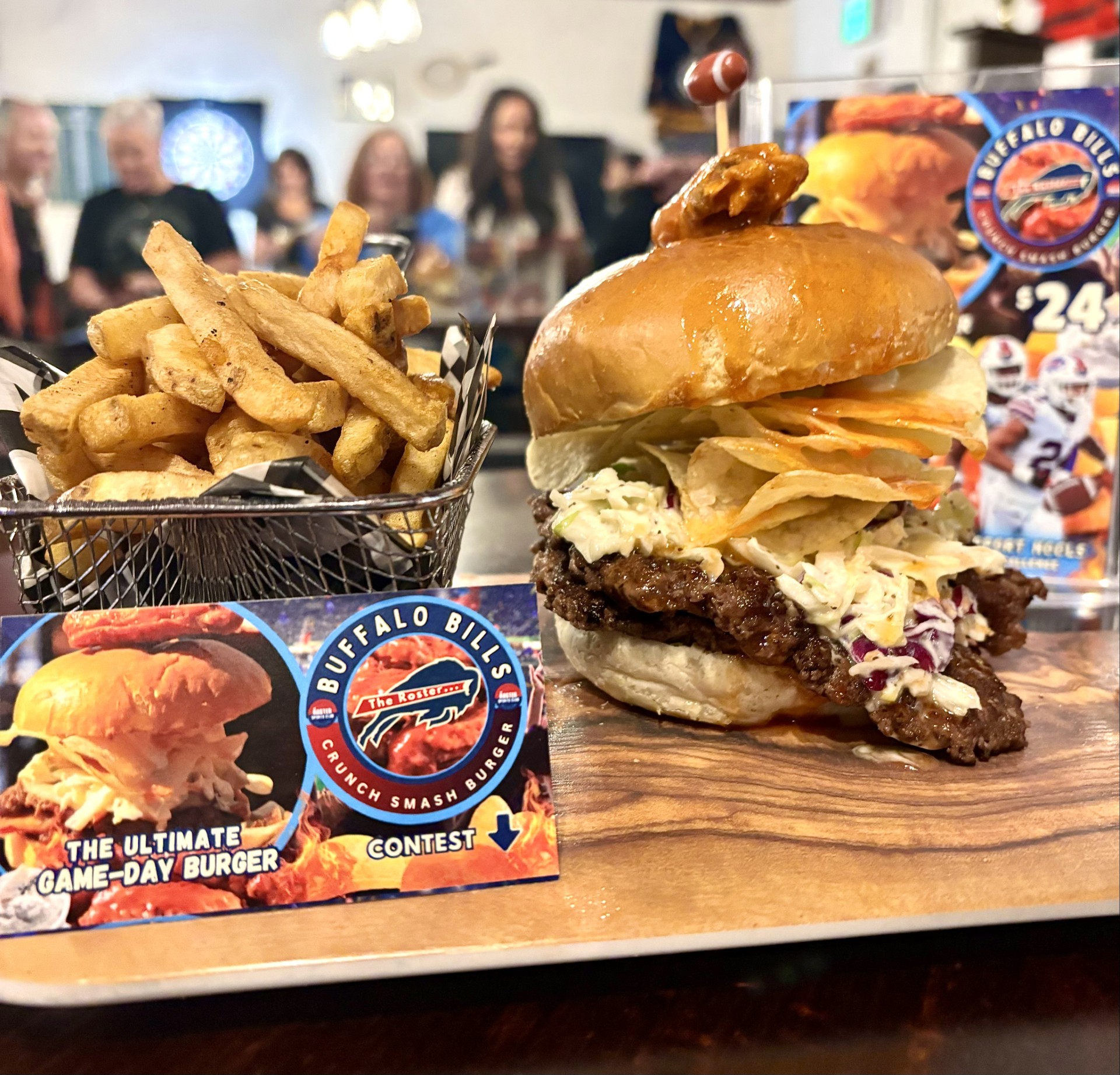 A beef burger with two beef smash patties, topped with mozza cheese, bacon, coleslaw, honey buffalo wing sauce, and potato chips on a Brioche bun with a mild blue-cheese aioli. Garnished on top with one seasoned chicken wing! A basket with french fries on the side.